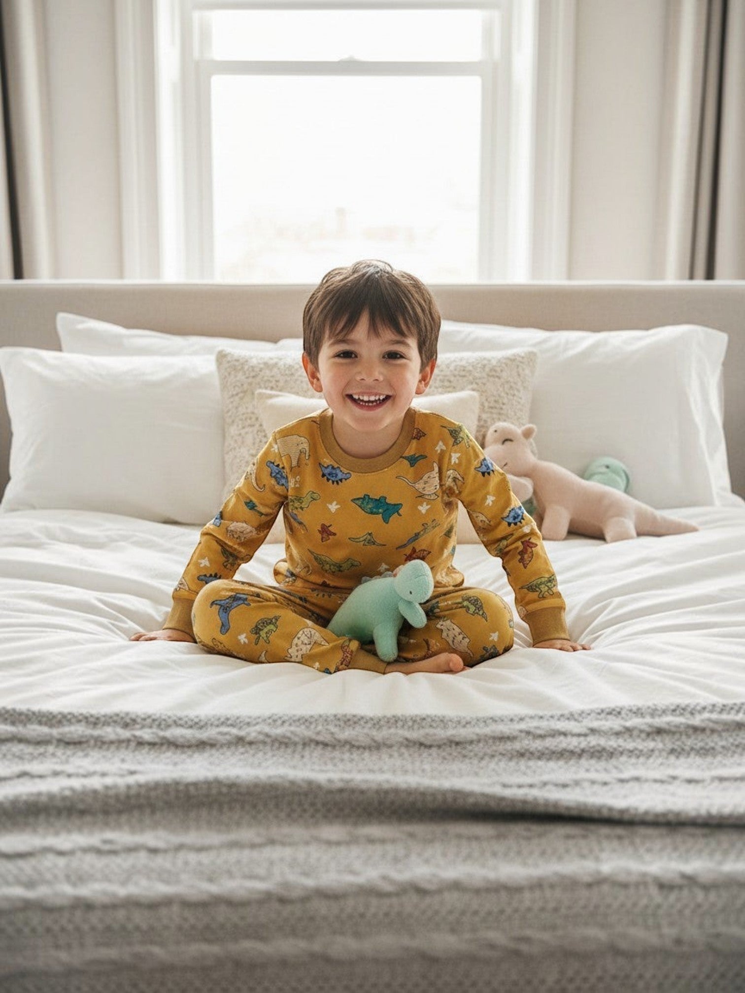 Niño sonriente sentado en la cama con pijama mostaza de Vamos Niños con estampado de dinosaurios y puños elásticos.