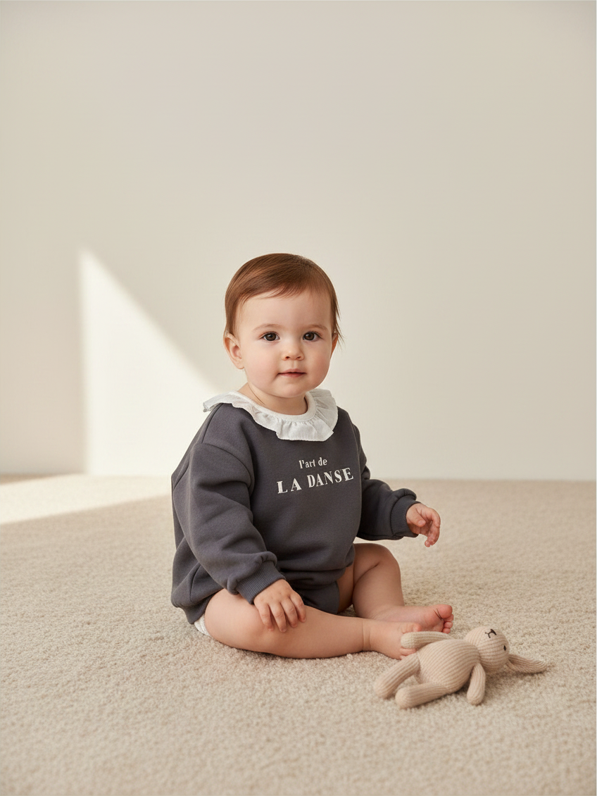 Bebé con polerón gris Lefties de Vamos Niños con cuello blanco y estampado, sentado sobre alfombra beige