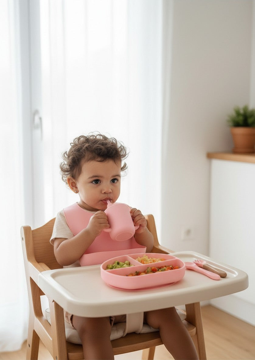 Bebé usando set de alimentación de silicona rosa Vamos Niños con plato, babero y vaso en trona de madera